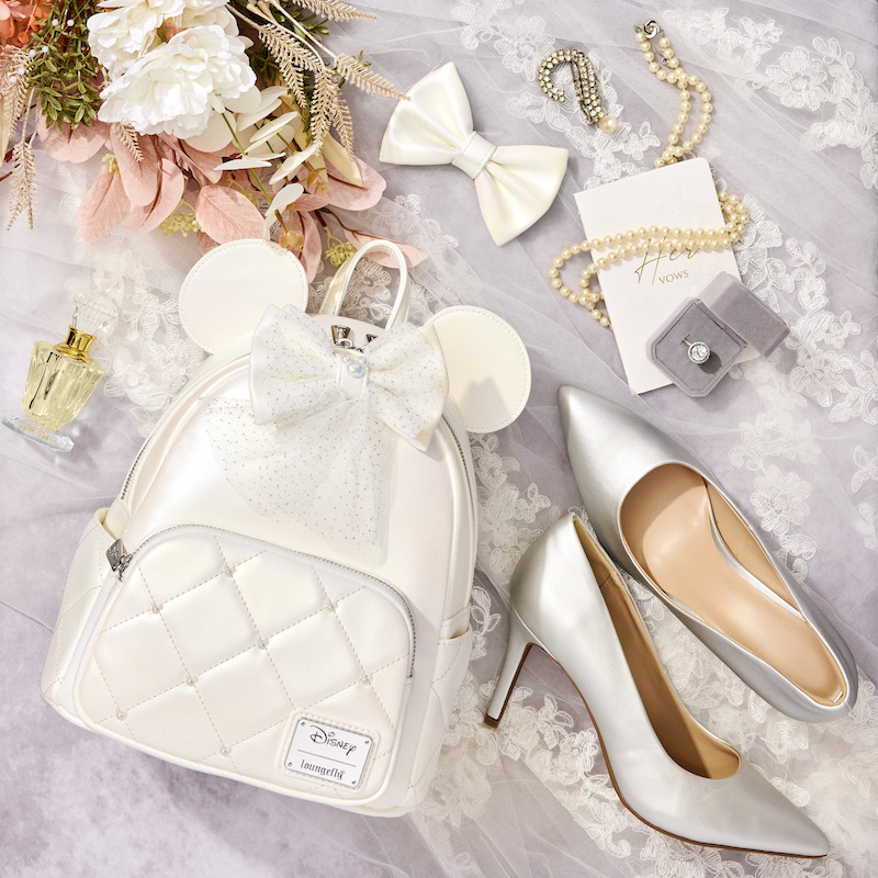 Loungefly Disney Minnie Mouse Iridescent Wedding Mini Backpack featuring a white iridescent mini backpack laying against a white tulle table covering and surrounded by various bridal accessories like silver high heels, a bow, a gold necklace, and flowers.